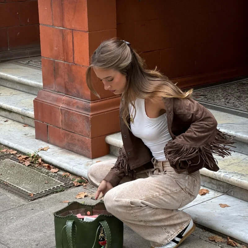 Woman in brown suede fringe jacket crouching with green bag on urban street.