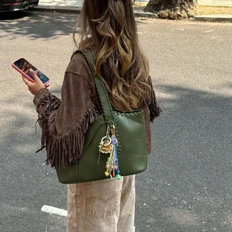 Woman in brown suede jacket holding a phone and an olive green bag, standing on a city street.