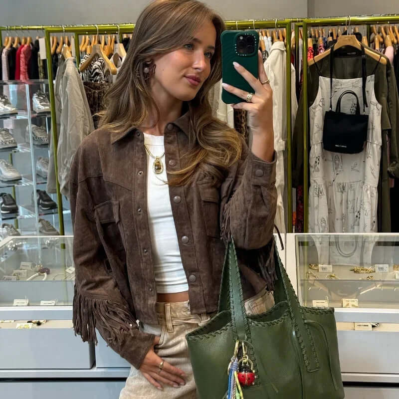 Young woman wearing a brown suede fringe jacket, styled with a white top and carrying a green handbag in a trendy retail store.