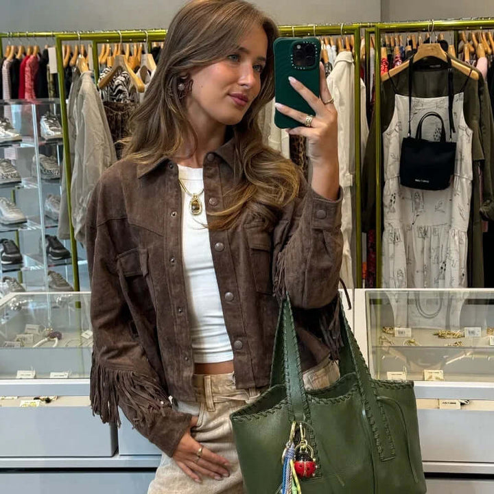 Young woman wearing a brown suede fringe jacket, styled with a white top and carrying a green handbag in a trendy retail store.