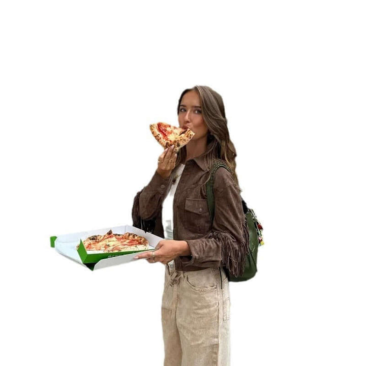 Young woman enjoying pizza while holding a box, wearing a brown suede jacket and light jeans.
